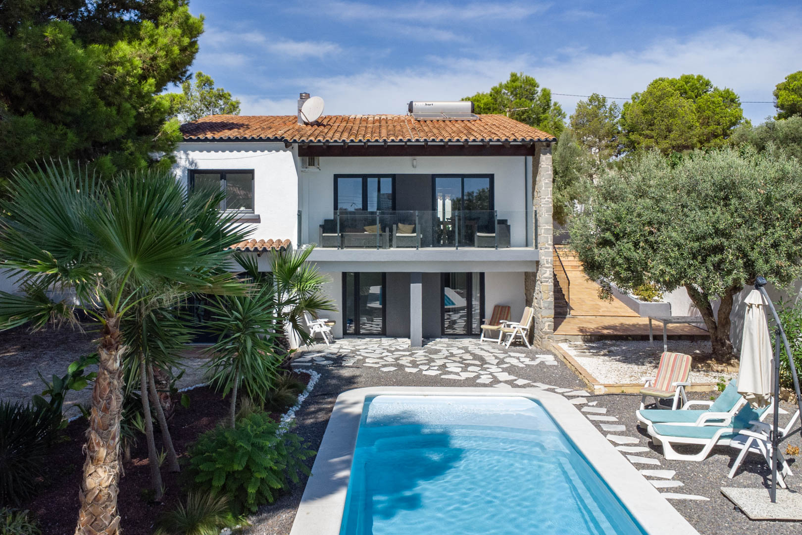 Exterior view of the villa featuring a rectangular pool, Mediterranean garden, and white facade. It includes glass-railed terraces and a solar water heater on the roof.