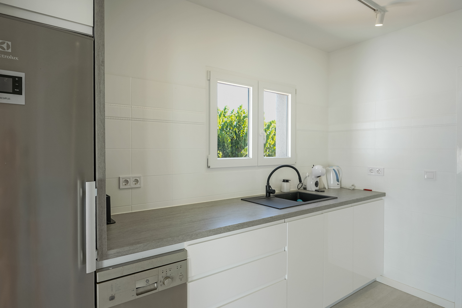 Functional kitchen featuring white handleless cabinetry, a grey countertop, black sink, and integrated appliances with natural light from a side window.