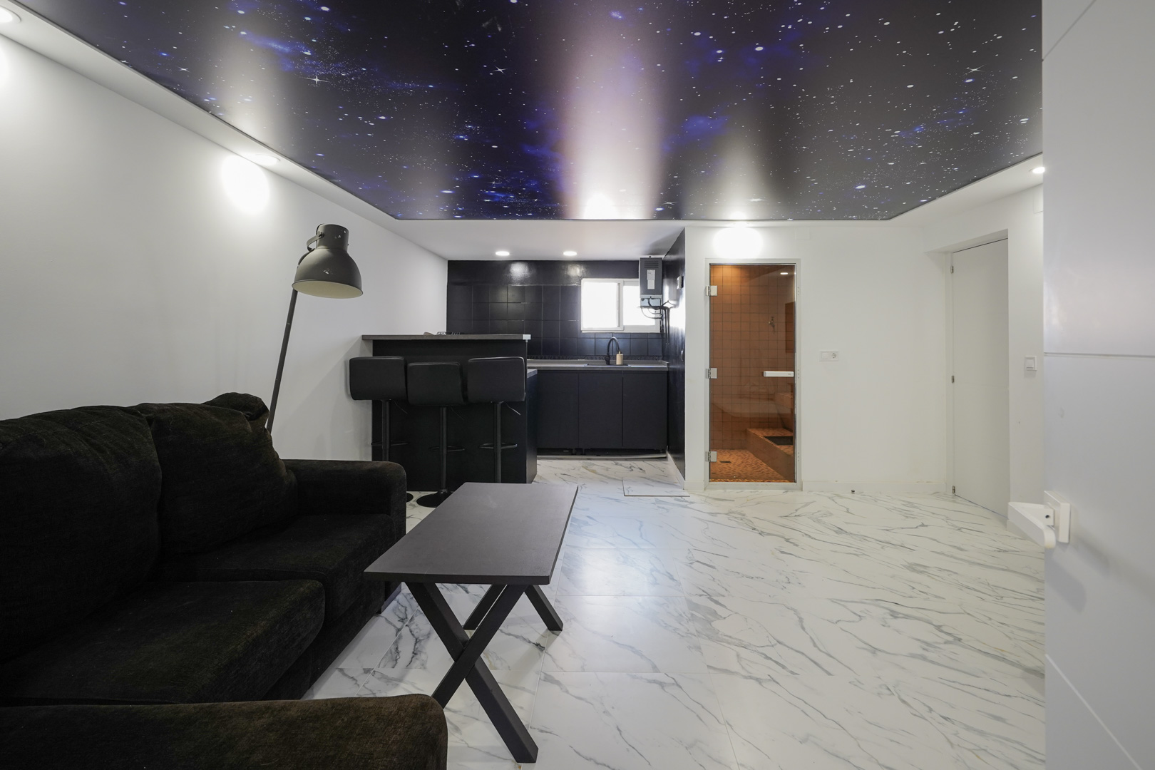 Living space with a modern kitchenette, marble-effect tiling, a distinctive galaxy-themed ceiling, and a private sauna behind a glass door.