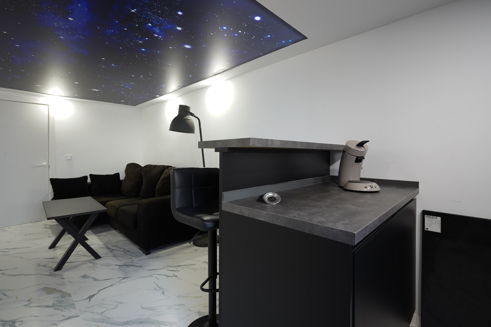 Lounge area featuring a coffee bar, dark sofa, and marble-effect flooring, characterized by a unique backlit ceiling panel with a galaxy motif.