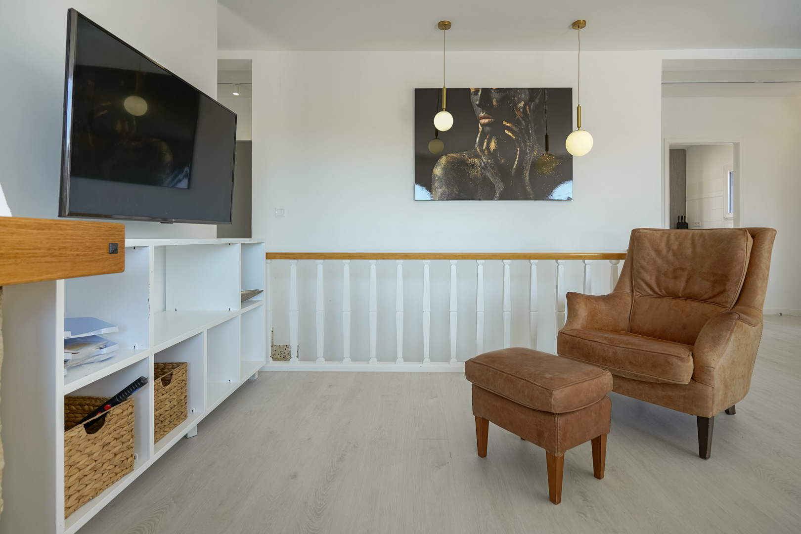 Lounge area featuring a leather armchair with ottoman, white shelving unit, and wooden balustrade. Modern globe pendant lights and light-toned flooring define the space.