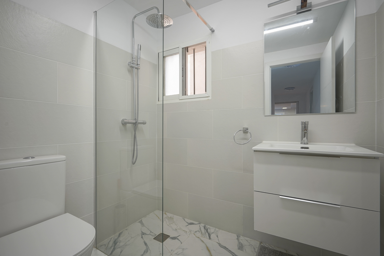 Modern bathroom featuring a walk-in shower with glass partition, rainfall showerhead, white vanity unit, and marble-effect floor tiles.