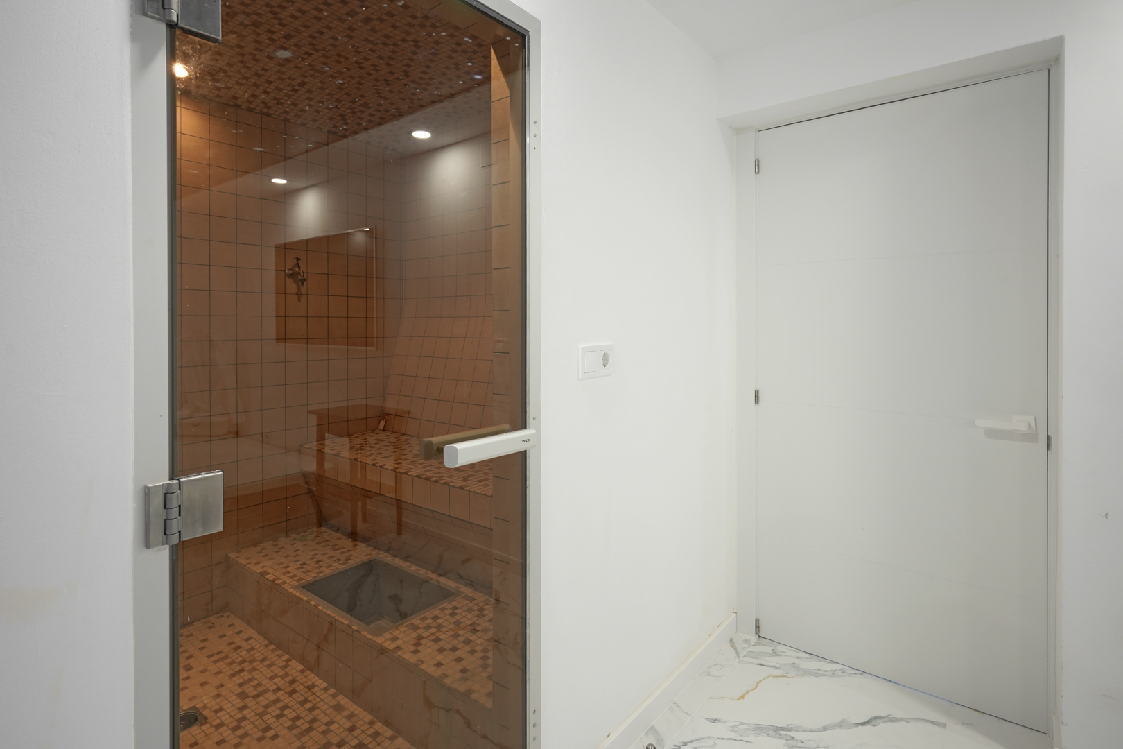 Private sauna featuring ceramic mosaic tiling, recessed ceiling lights, and a glass door, located adjacent to a room with marble-effect flooring.