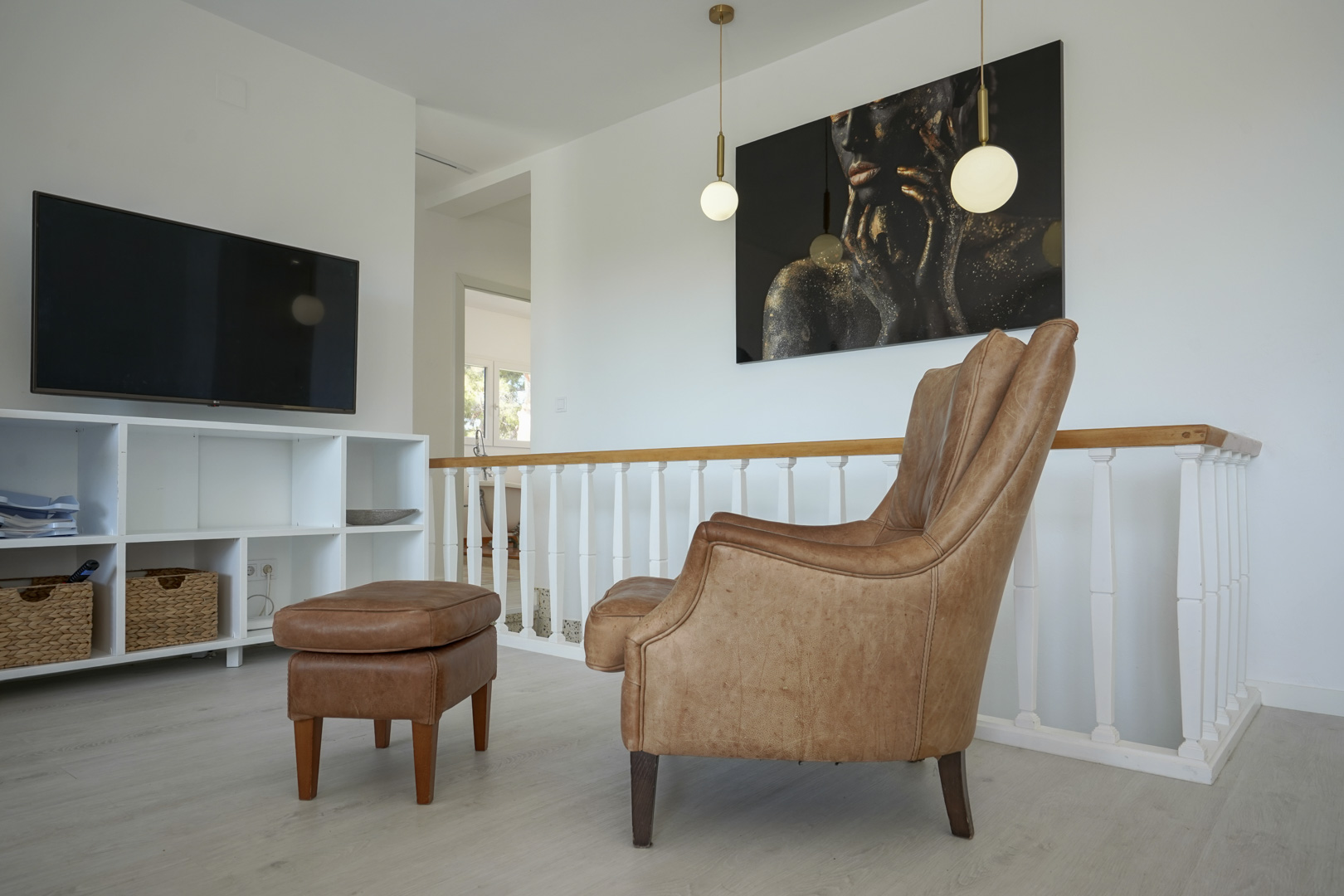 Reading nook with a leather armchair and ottoman next to the staircase railing. Features a wall-mounted TV, white shelving, and modern pendant lighting.