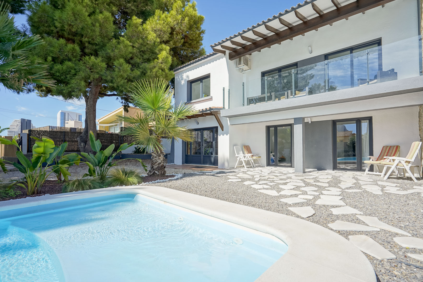 Rear facade featuring a swimming pool, upper terrace with glass railings and wooden beams, and a gravel patio with irregular white stepping stones.