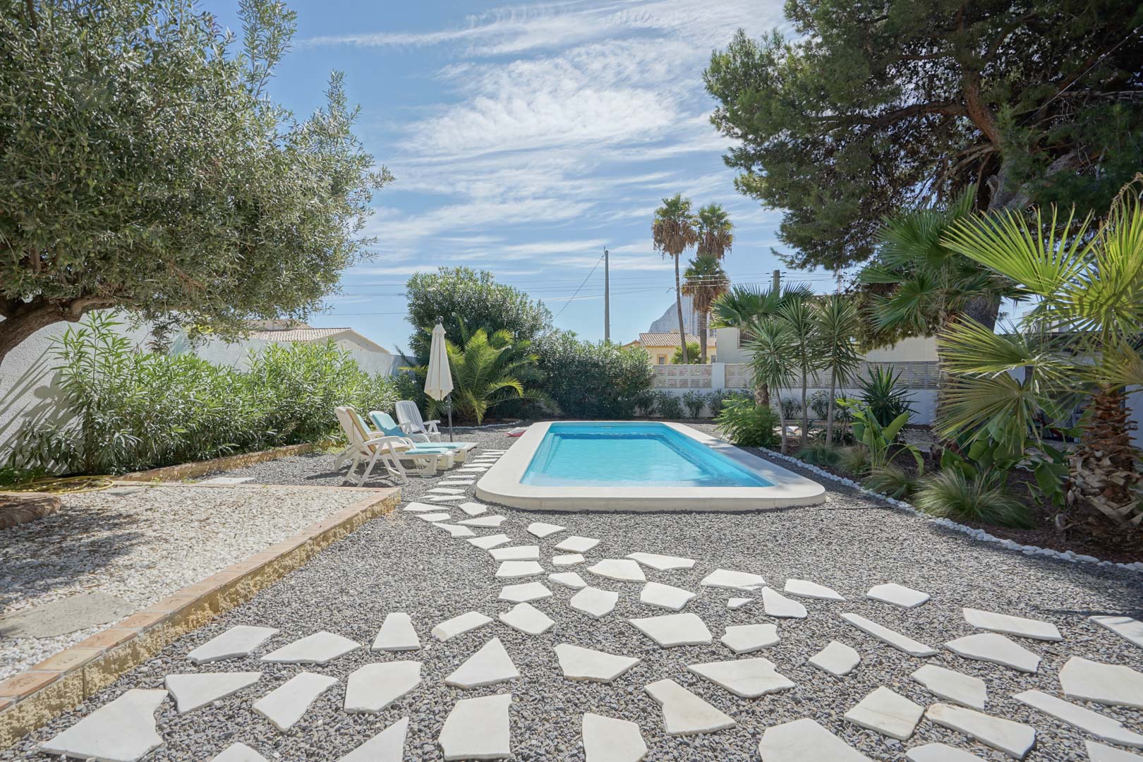 Rectangular swimming pool set within a Mediterranean garden featuring olive trees, palms, and a decorative stone pathway over grey gravel.