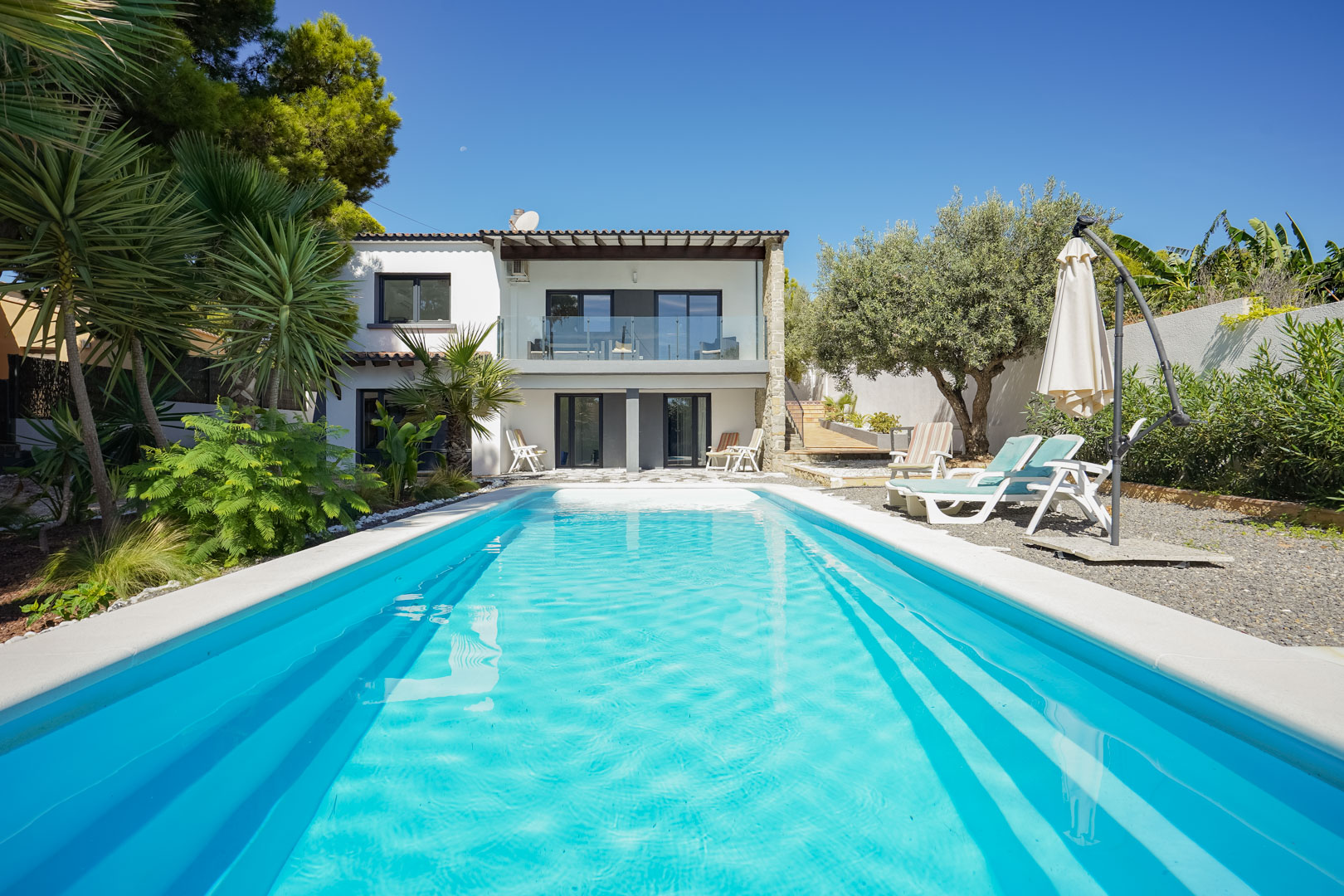 View of the rectangular swimming pool and Mediterranean garden with palms, showing the villa's white facade and upper terrace with glass railings.