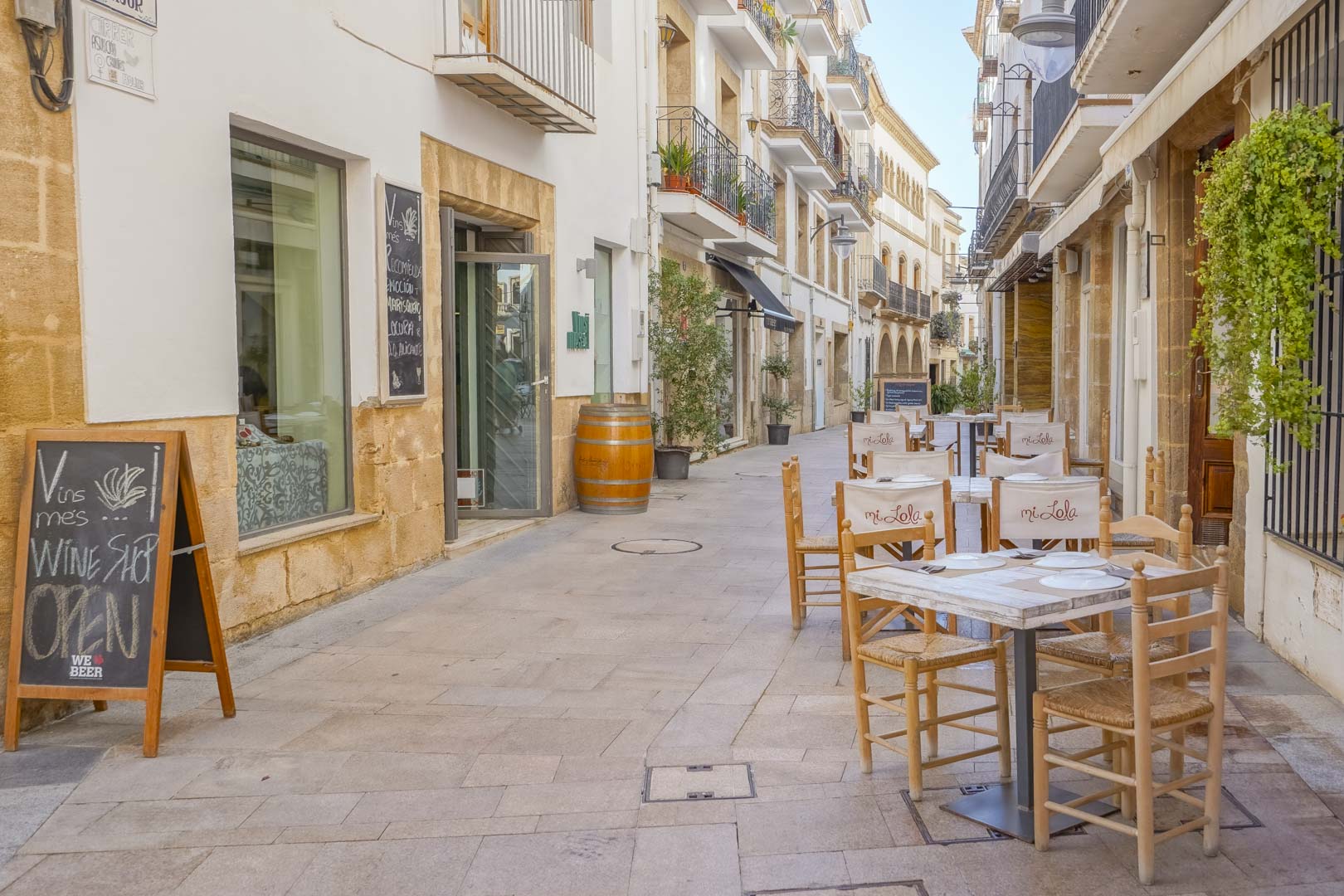 Pedestrian street in Jávea's historic center featuring traditional stone facades, wrought iron balconies, and local restaurant terraces near the new development.