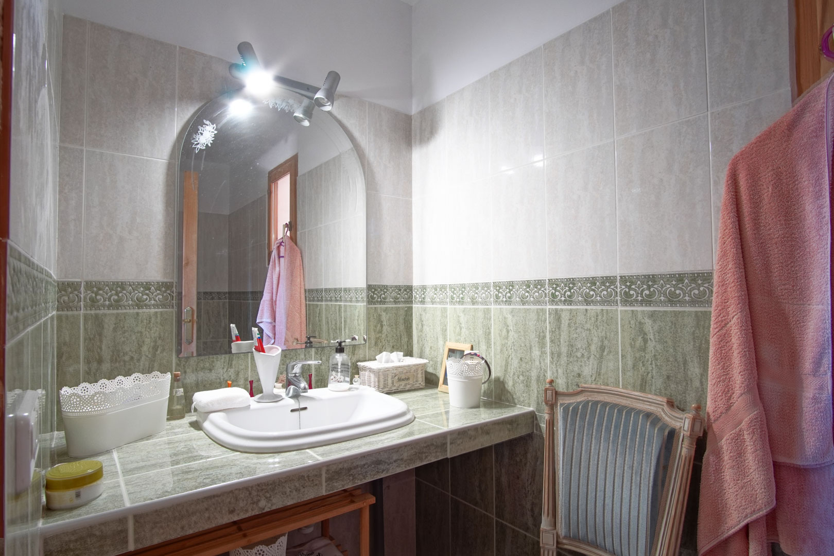 Bathroom featuring a green tiled countertop with an inset sink, an arched mirror with overhead spotlights, and wall tiles with a decorative border.