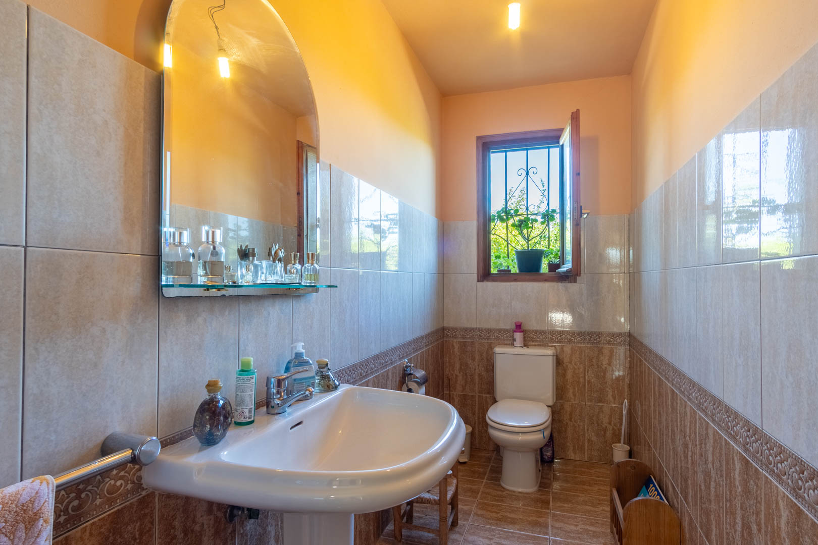 Bathroom featuring earth-toned wall tiles, a pedestal sink with an arched mirror, and a wooden window providing natural light.