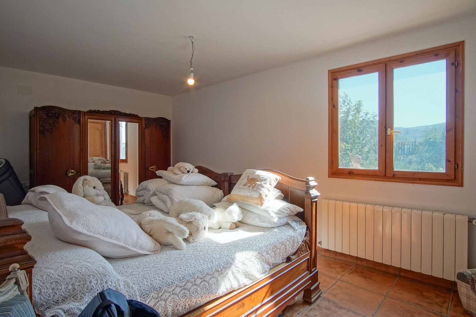 Bedroom featuring carved wooden furniture, a mirrored wardrobe, terracotta tile flooring, and a window overlooking the natural landscape with a radiator.