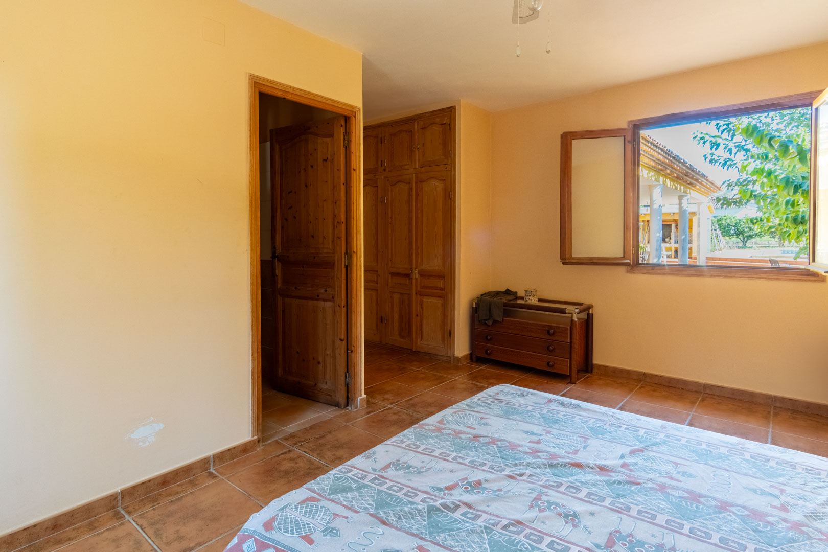 Bedroom featuring rustic terracotta flooring, built-in wooden wardrobes, and a window overlooking the exterior porch and garden area.