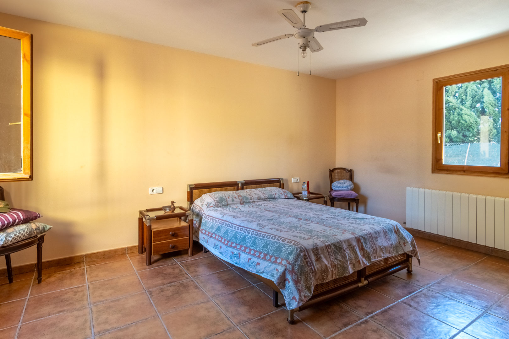 Bedroom featuring terracotta tile flooring, warm-toned walls, and a white ceiling fan. The space includes wooden-framed windows, a radiator, and rustic-style wooden furniture.