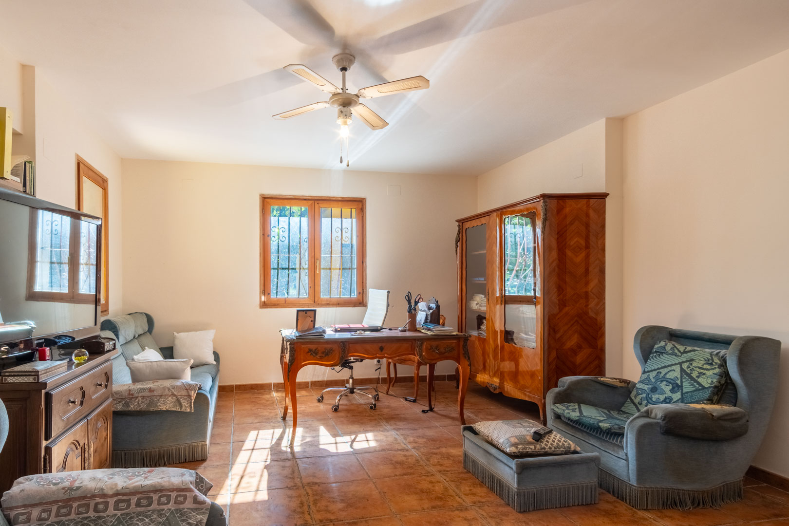 Bright home office featuring terracotta tile flooring, a classic wooden desk, and matching armoire. The space includes upholstered seating and a ceiling fan.