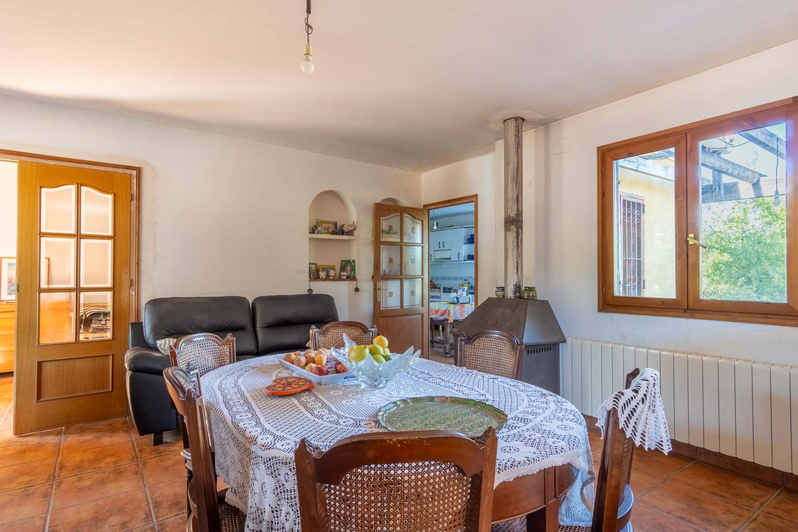 Dining area featuring terracotta floors, a wood-burning stove, and a classic wooden table set. Natural light enters through a timber-framed window, illuminating the interior.