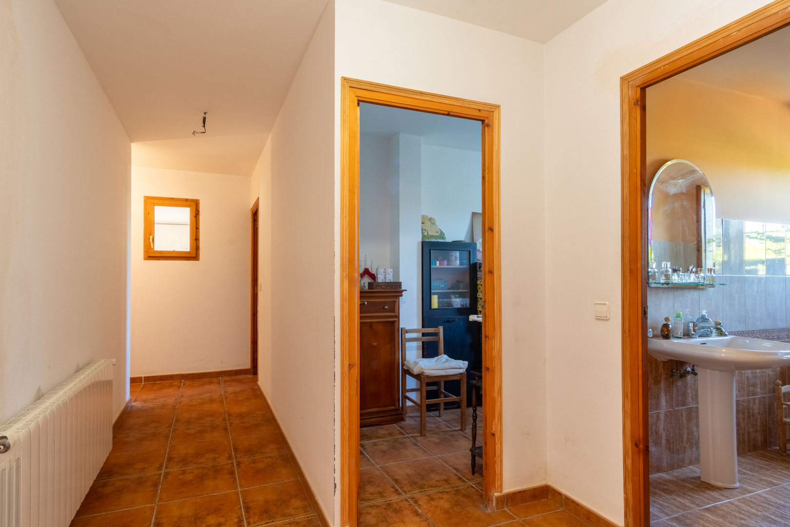 Distribution hallway featuring rustic tiled flooring and wooden frames, providing access to a bathroom with a pedestal sink and rooms with radiator heating.