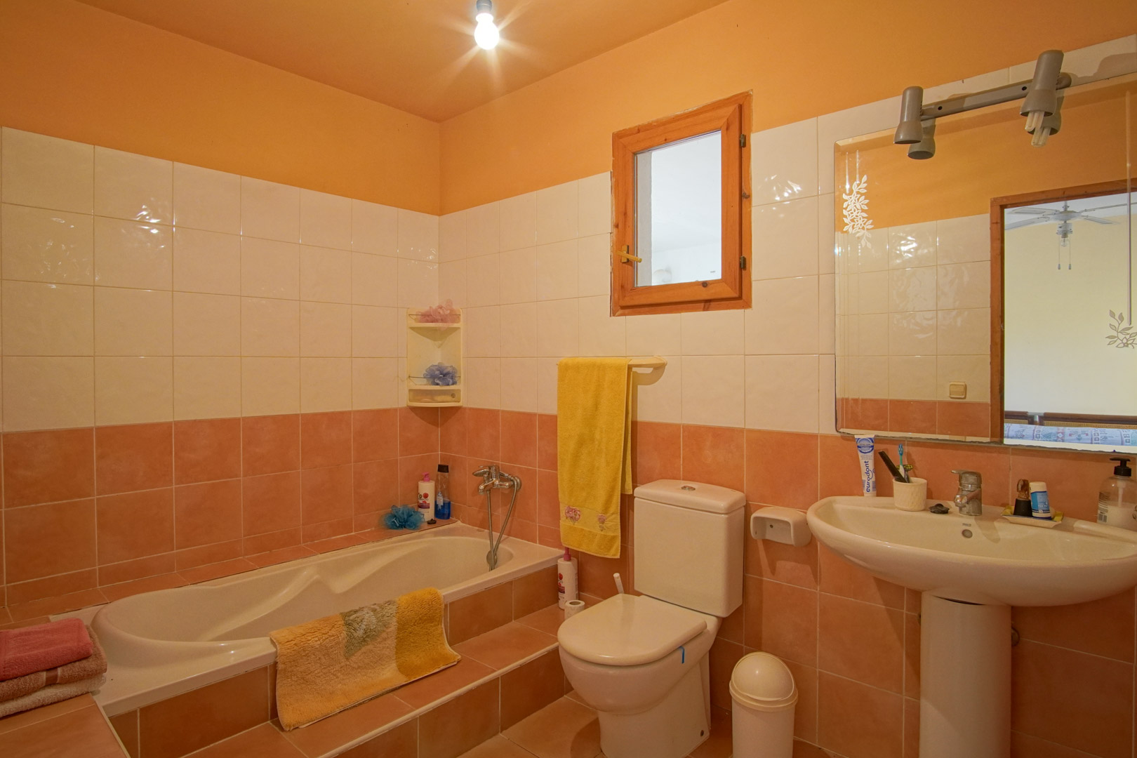 Functional bathroom with salmon and white tiling, a built-in bathtub with an access step, a pedestal sink, and a small window with a natural wood frame.