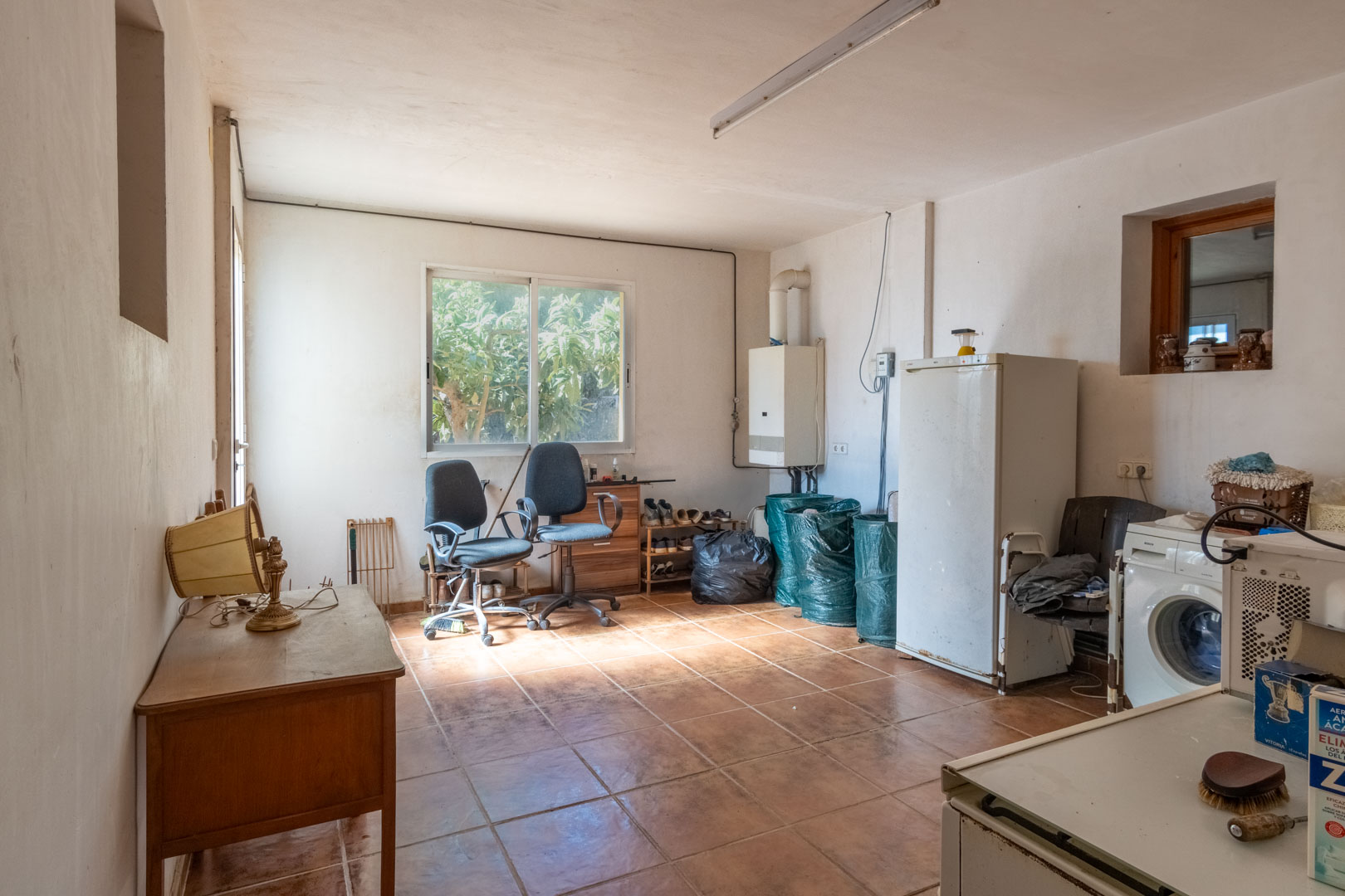 Functional laundry and storage room featuring terracotta tile flooring, a washing machine, wall-mounted boiler, and natural light from a garden-facing window.