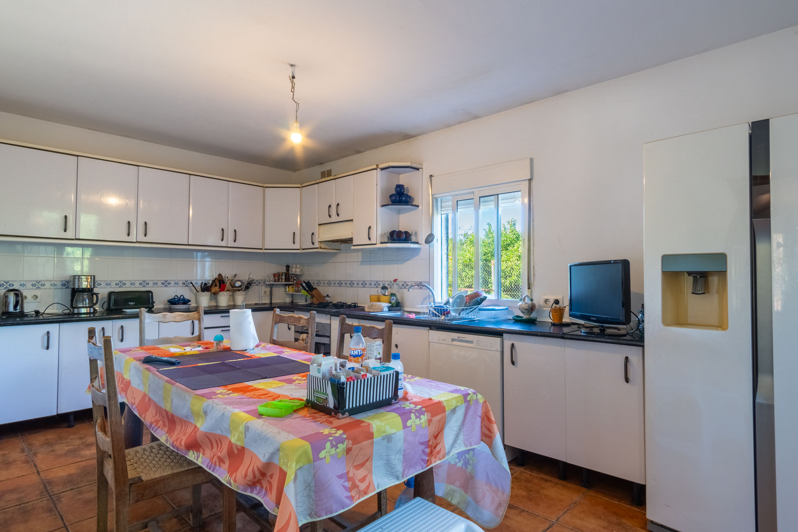 Independent kitchen with white cabinetry, dark countertops, and a central dining area. Features ceramic tile flooring and a window overlooking the garden.