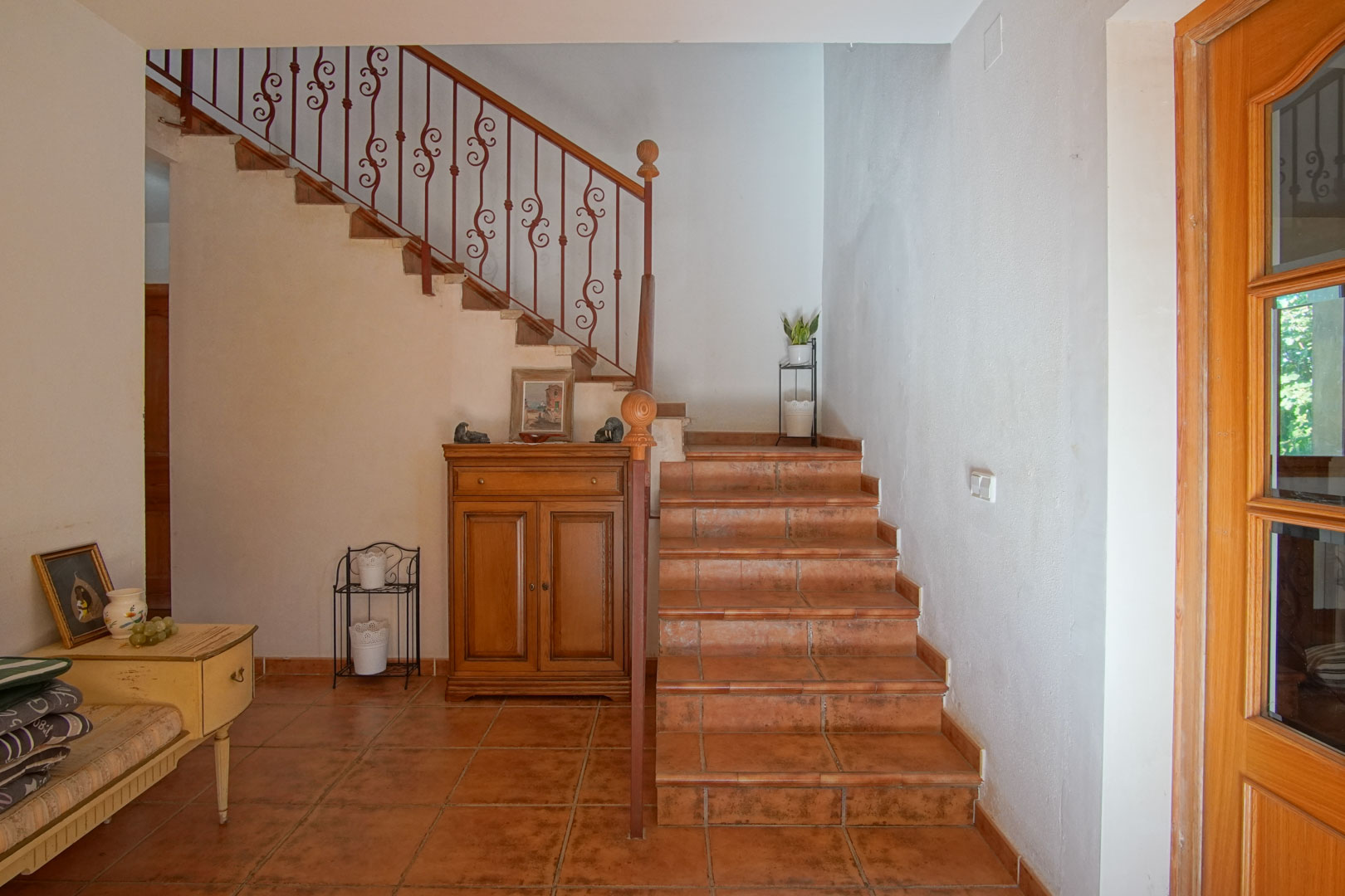 Interior staircase featuring rustic terracotta tiles and an ornate wrought iron railing. The hallway includes matching clay floors and traditional wooden furniture.
