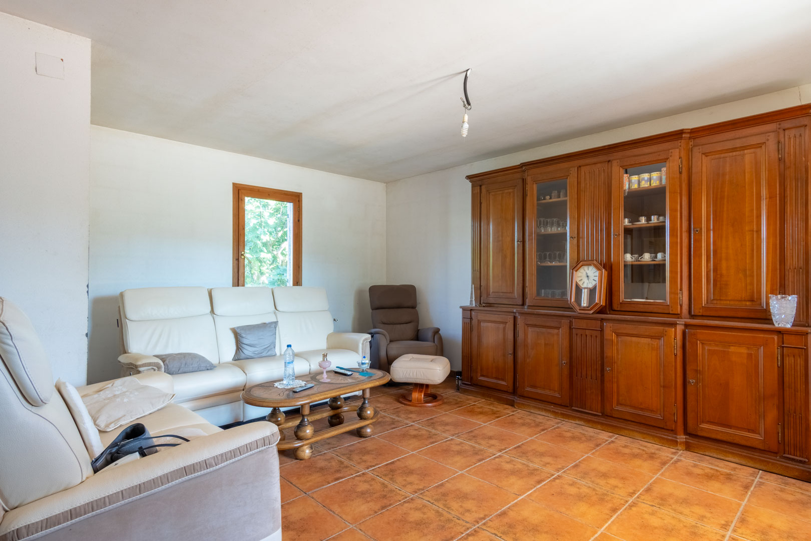 Living area featuring terracotta tile flooring, white leather sofas, and a large solid wood wall unit with glass display cases.