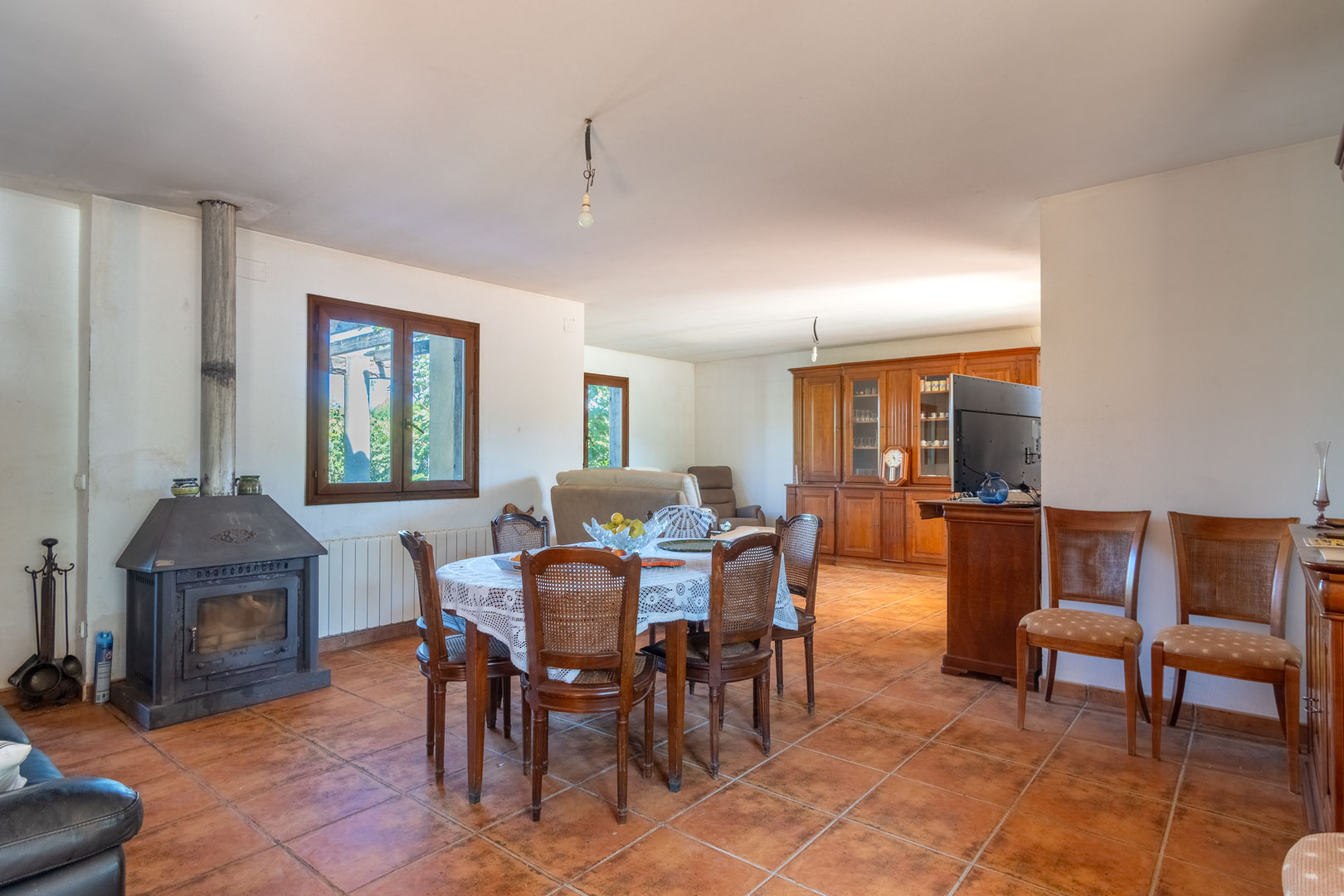 Living-dining area featuring rustic tile flooring, a traditional wood-burning stove, and classic wooden furniture including a large display cabinet.