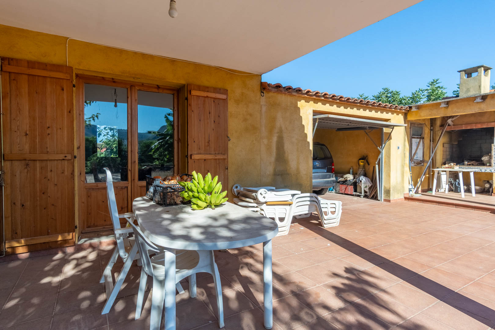 Outdoor terrace featuring terracotta tiling, a shaded dining area, and access to a covered parking space and traditional BBQ kitchen.
