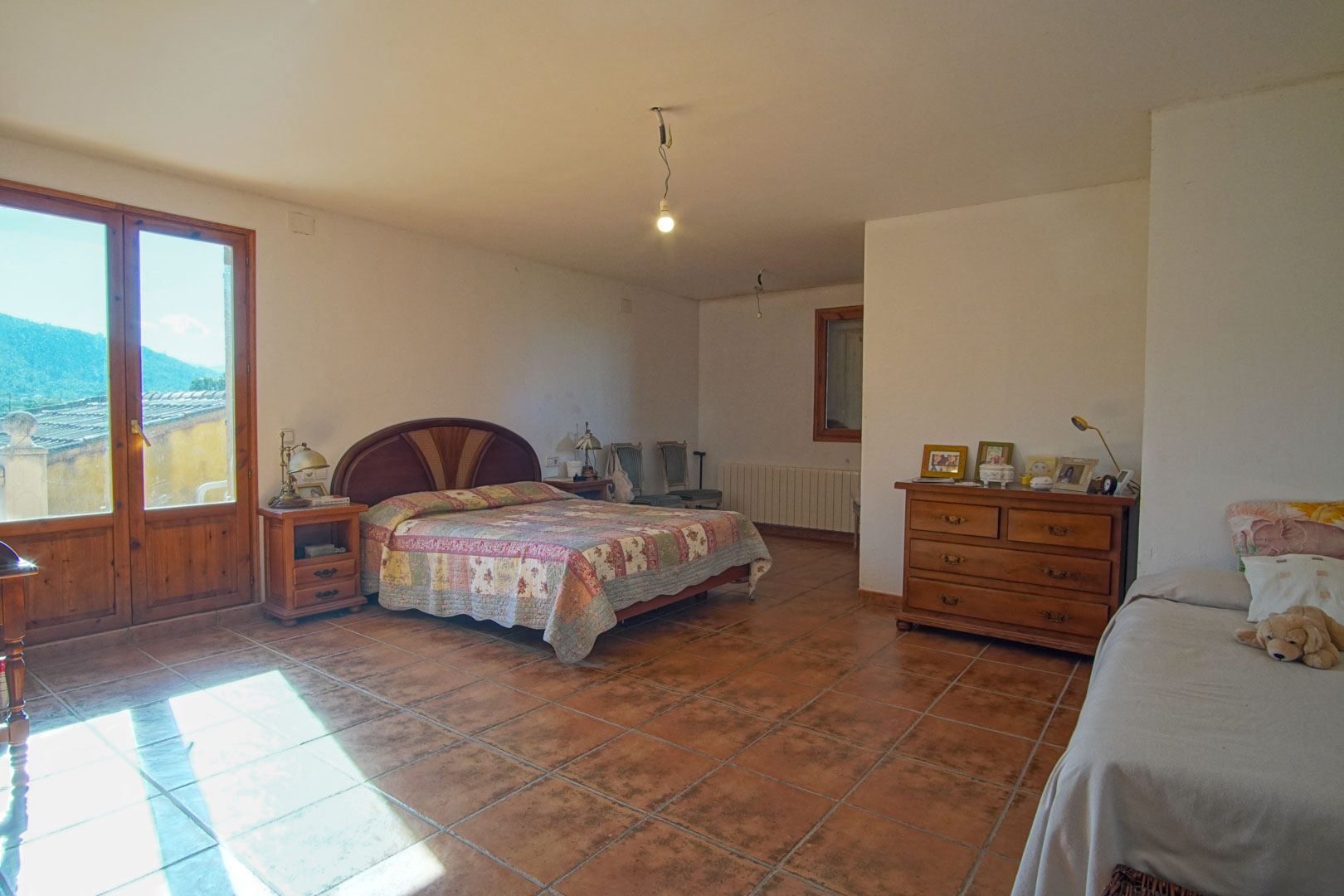 Spacious bedroom featuring rustic terracotta flooring, classic wooden furniture, and a glass door providing natural light and mountain views.