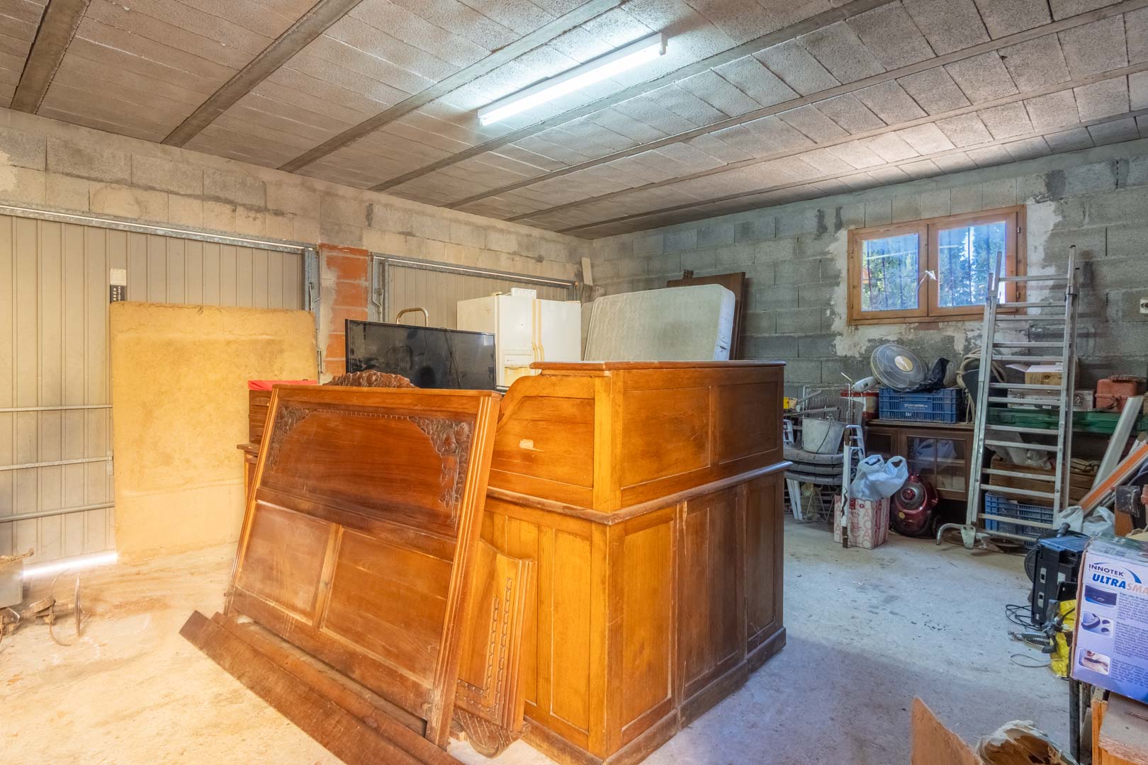 Spacious storage area or garage featuring concrete block walls, exposed ceiling beams, fluorescent lighting, and a wooden-framed window for natural light.