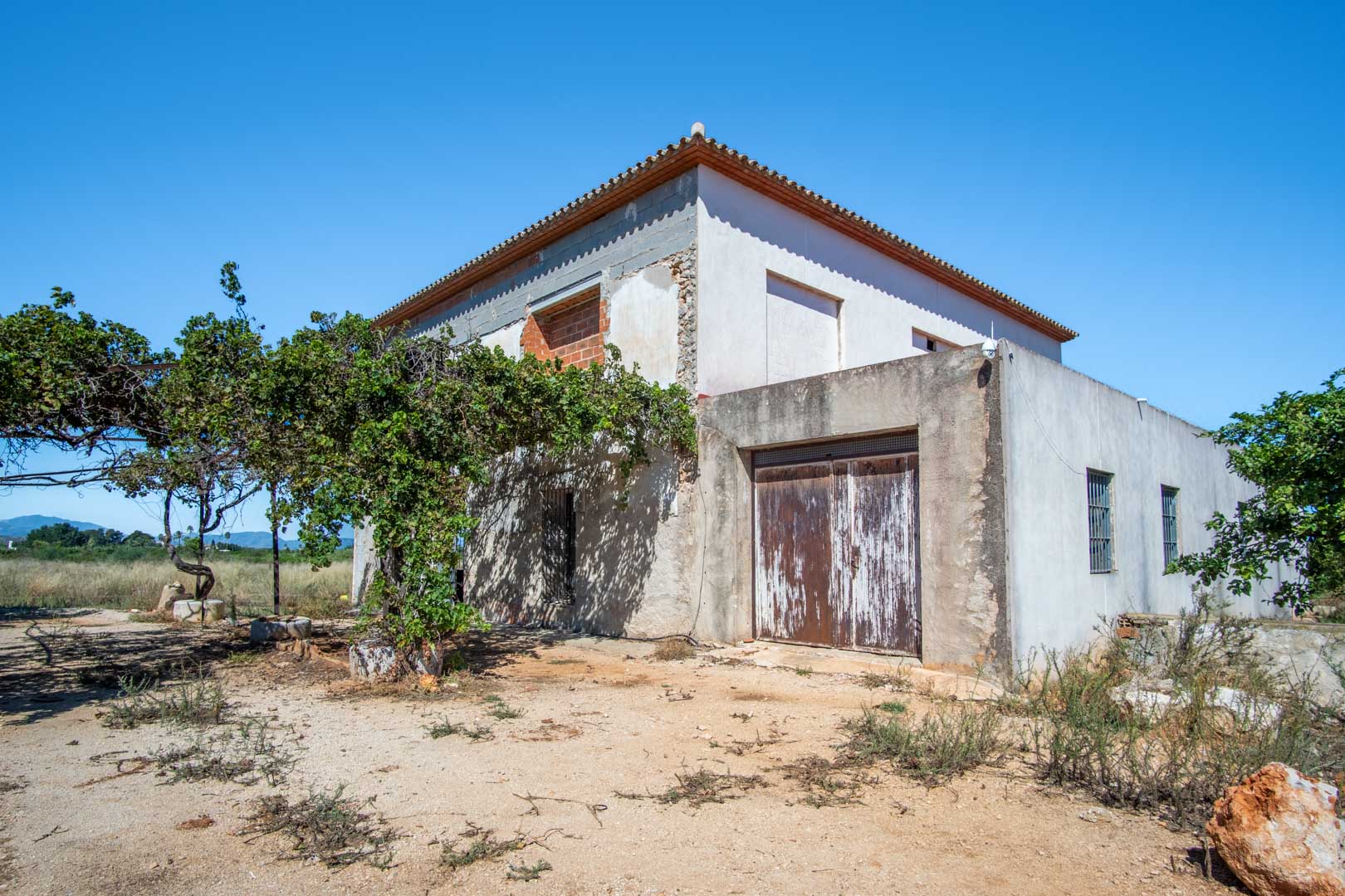 Exterior view of the rural house under renovation, featuring a consolidated structure, flat plot, and open views towards the surrounding mountains.