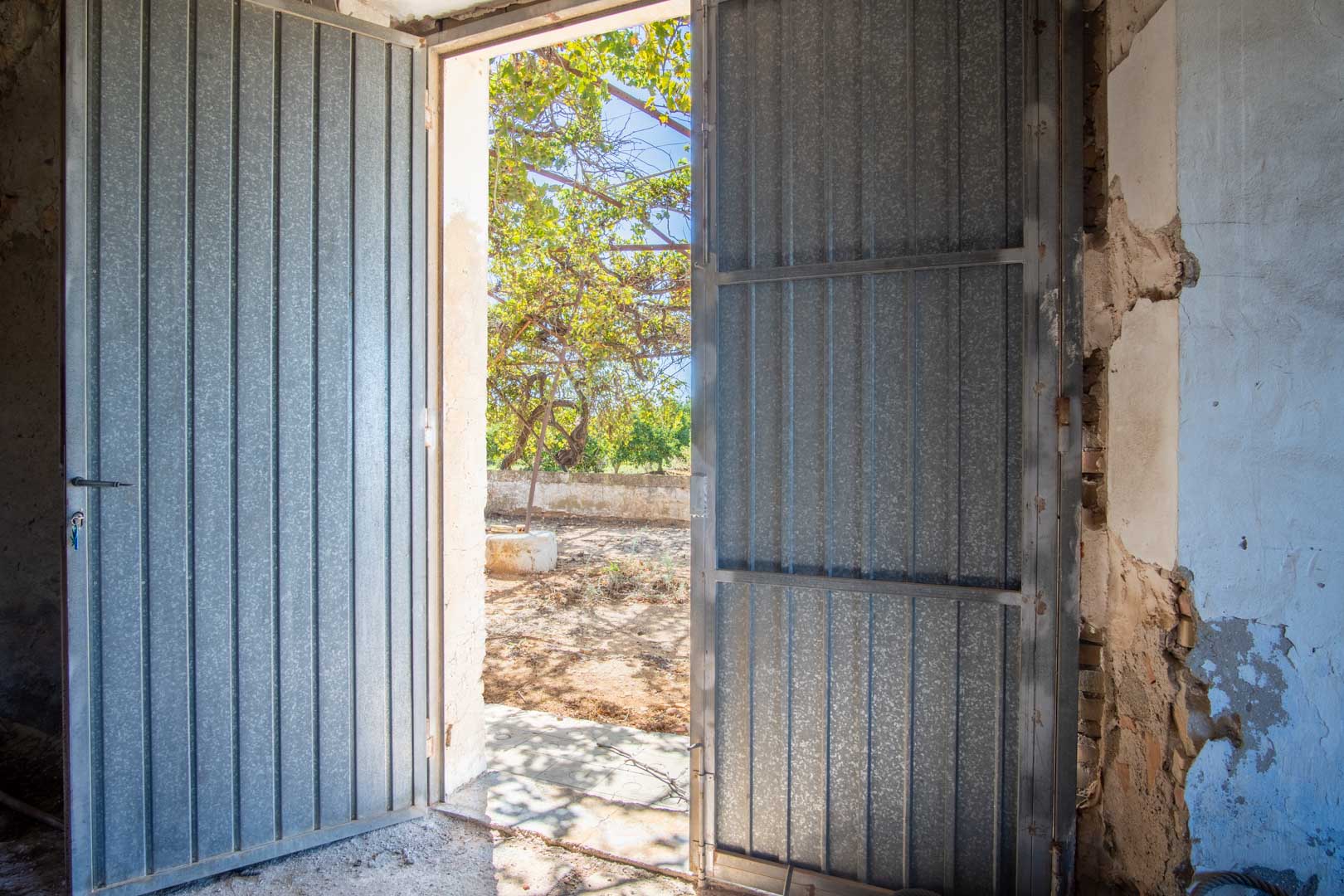Main access through large metal doors connecting the open-plan interior with the outdoor plot and its Mediterranean vegetation.