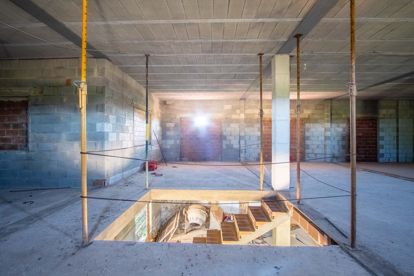 Open-plan interior under construction featuring concrete block walls, steel beams, and a floor opening for a staircase to the lower level.
