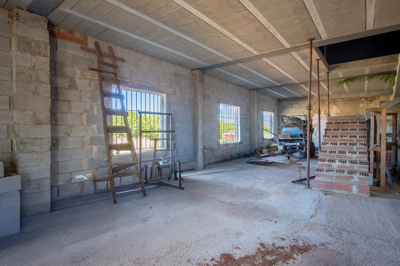 Spacious open-plan interior under renovation featuring concrete block walls, a beam-and-block ceiling, and windows with security bars offering natural light.