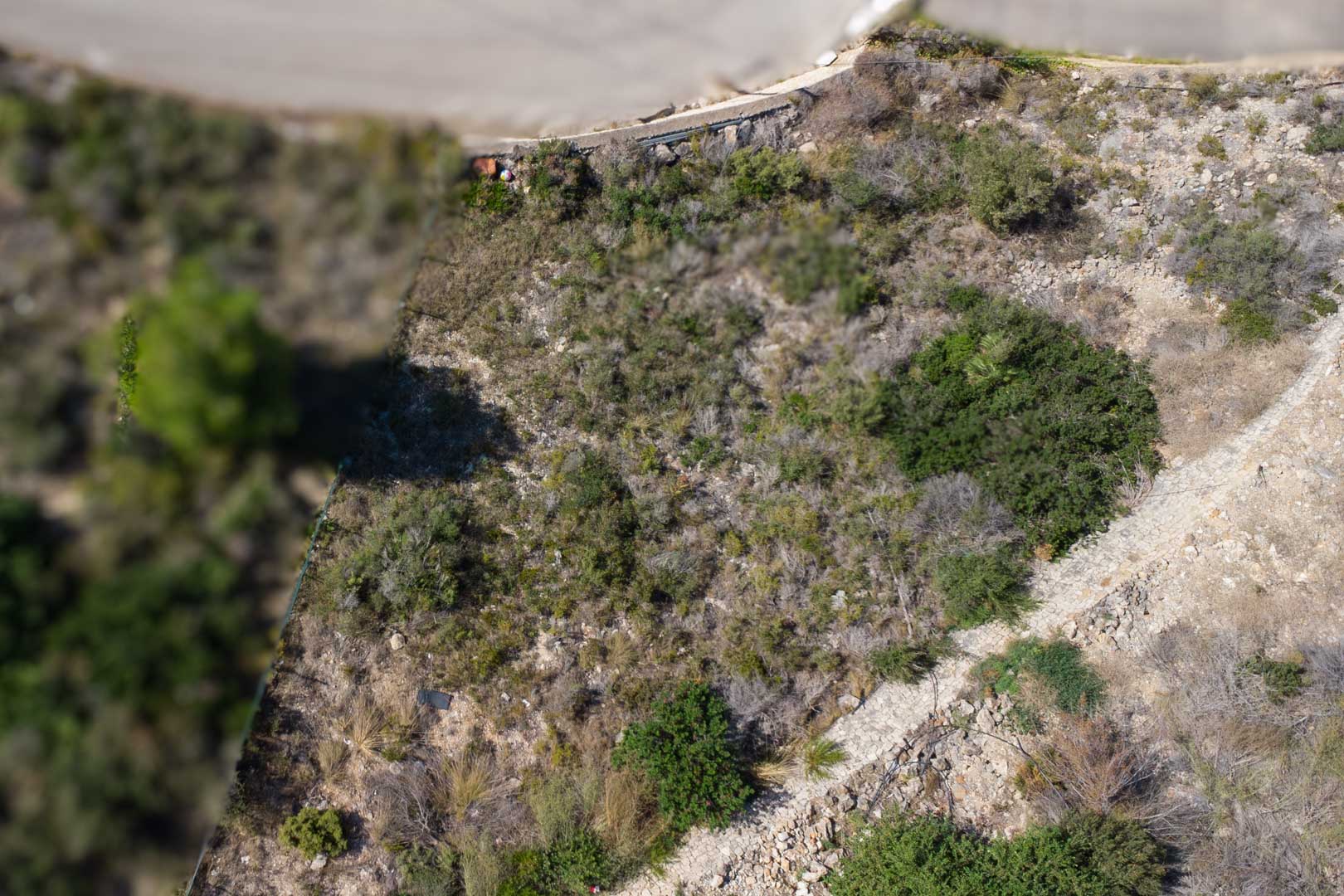 Aerial view of the sloping urban plot featuring native Mediterranean vegetation, rocky terrain, and a stone boundary path.
