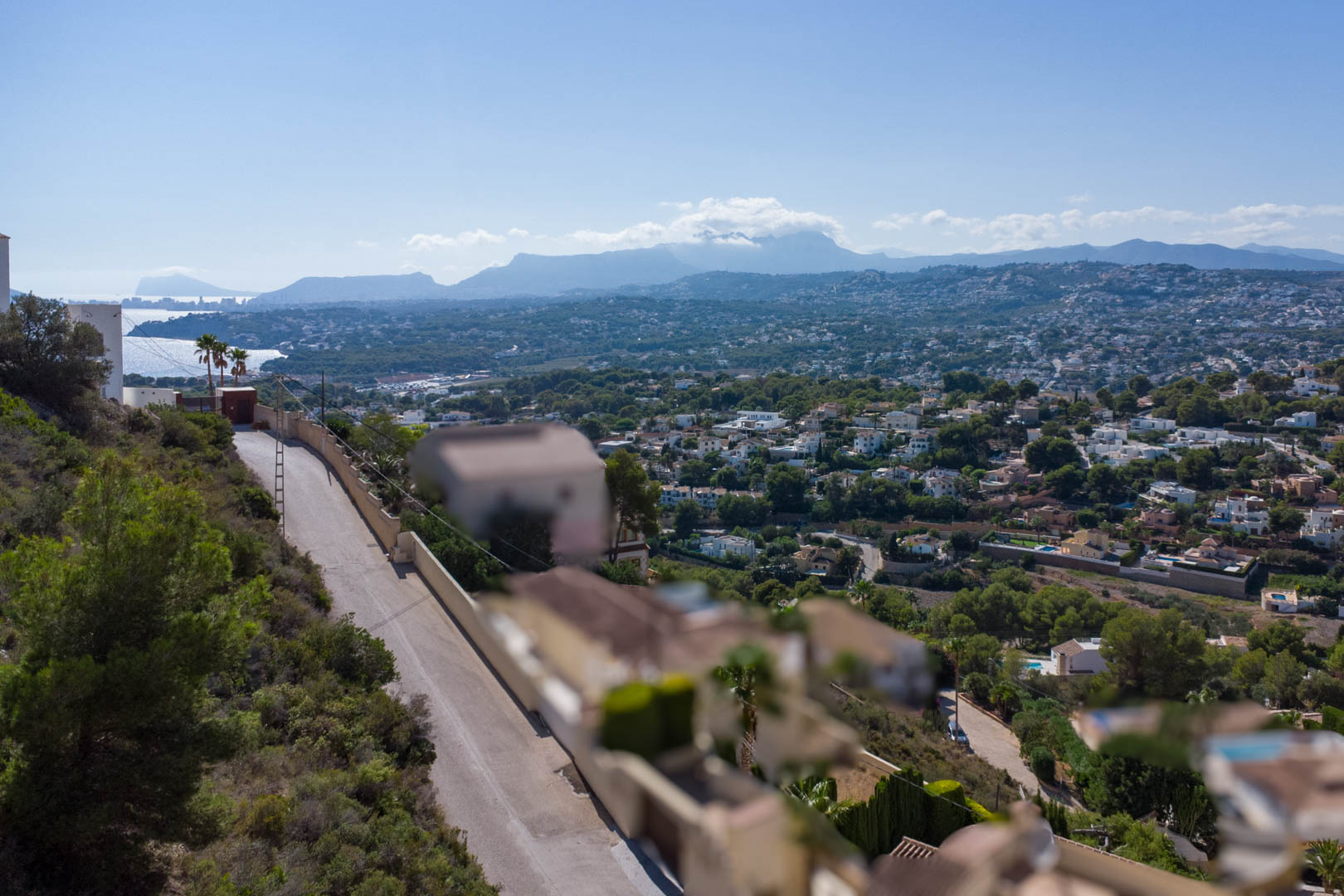Panoramic view from the elevated plot in El Portet, overlooking the residential area, the Moraira coastline, and distant mountain ranges under a clear sky.