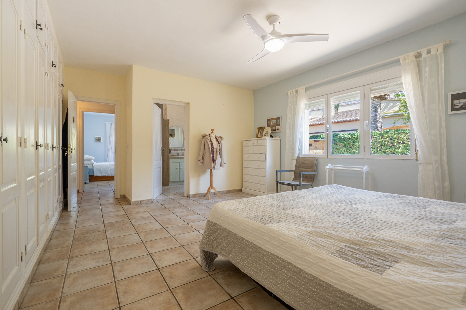 Bright bedroom with built-in wardrobes, ceiling fan, and en-suite bathroom. Cream-colored tile flooring.