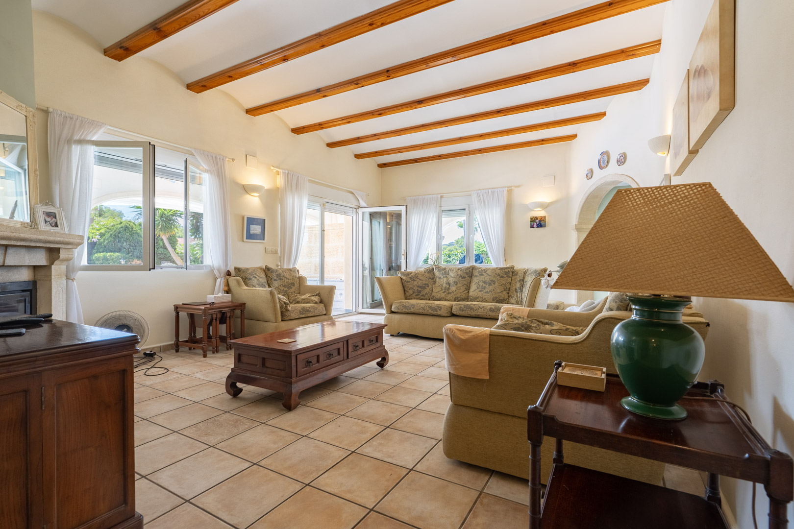 Bright living room with wood beams, patterned sofas, and tile flooring. Windows feature white curtains.