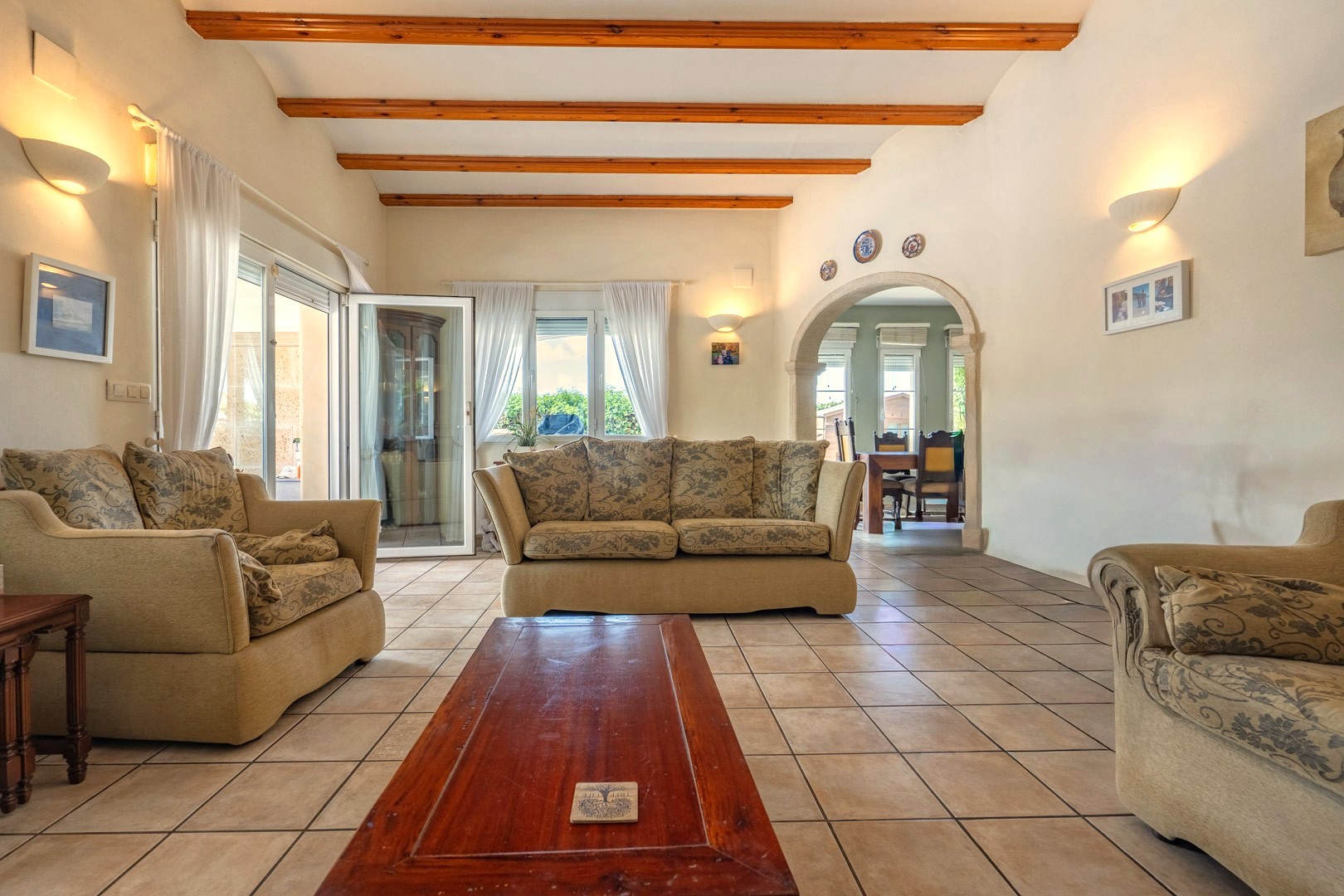 Bright living room with wood beams, patterned sofas, and tiled floor. Access to dining room.