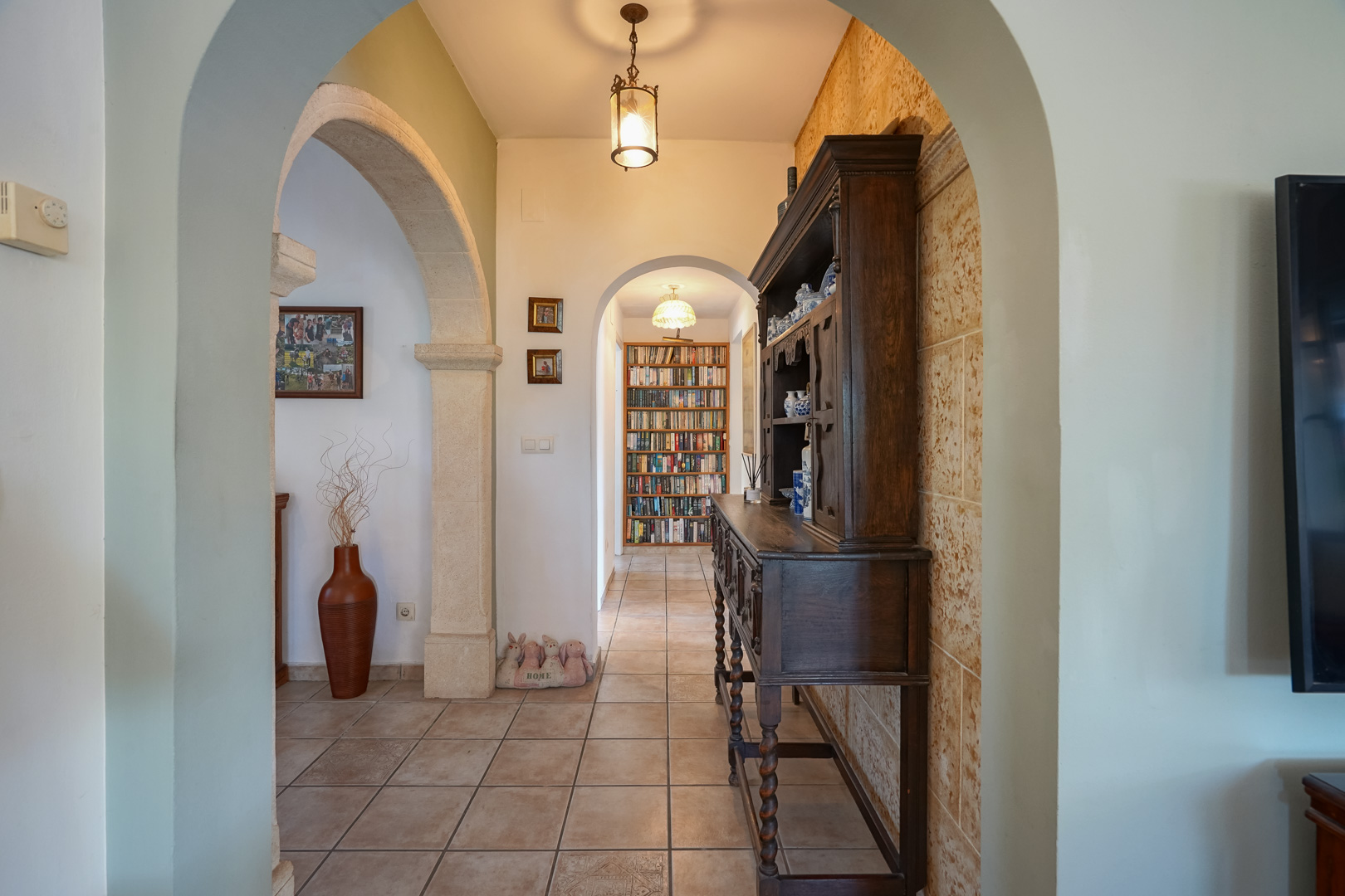 Hallway with stone arches and a library in the background. Dark wood furniture.