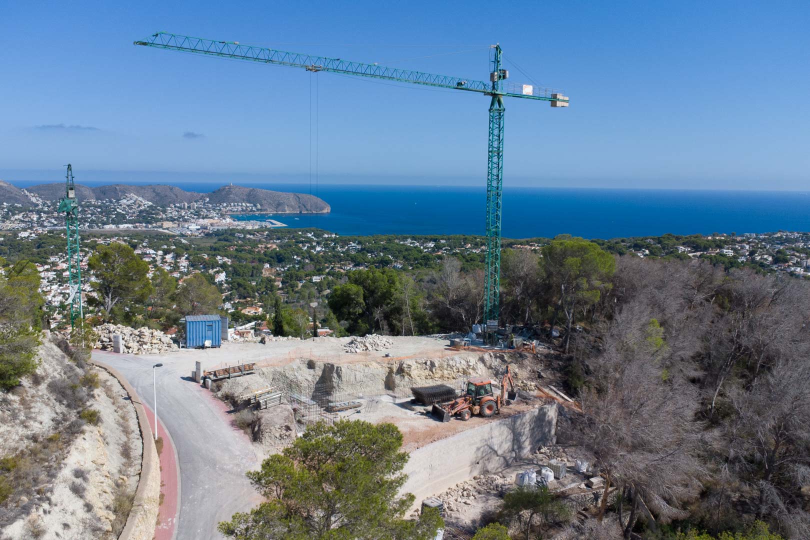 Elevated view of the construction site featuring a tower crane and excavation works on a hillside plot overlooking the Mediterranean coastline in Moraira.