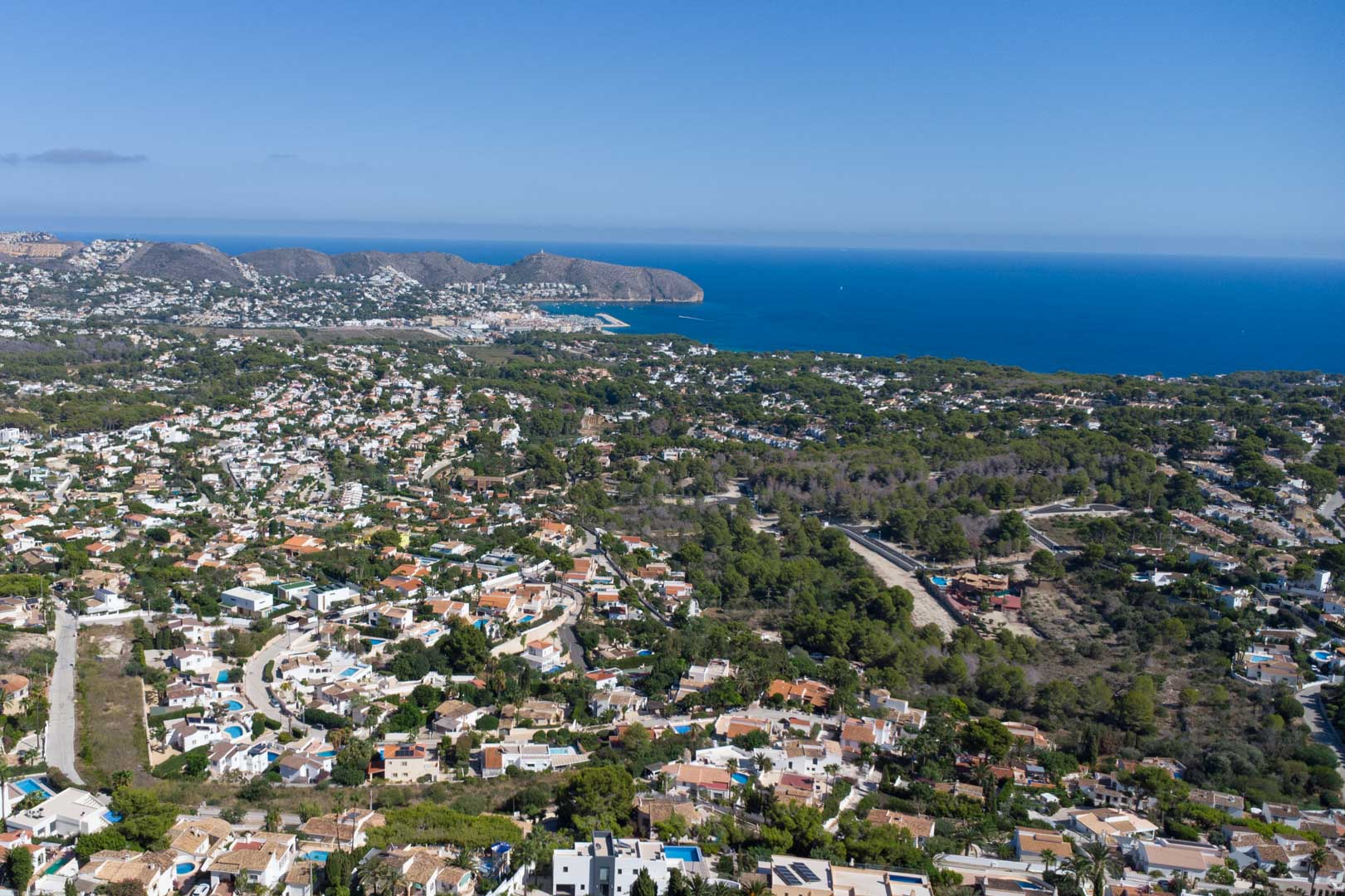 Panoramic aerial view of Moraira's coastline, featuring the marina and Cap d'Or headland, with residential villas nestled among Mediterranean pine groves.