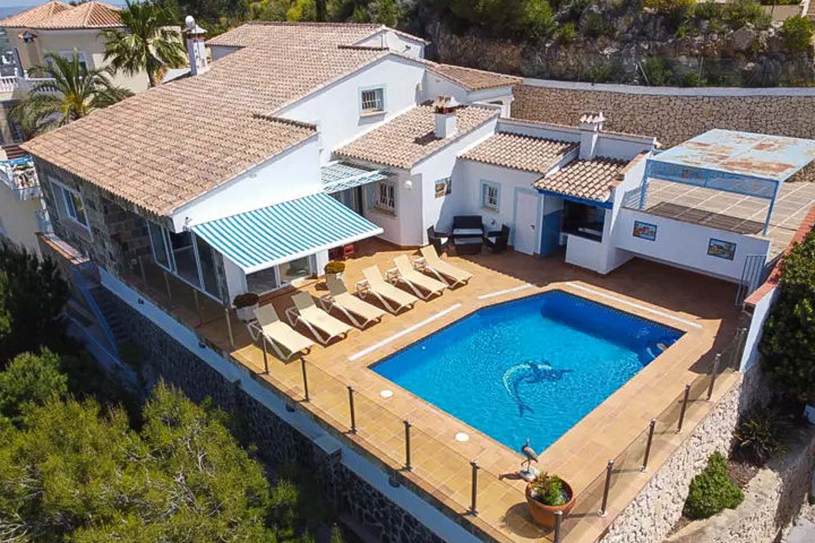 Aerial perspective of the villa showing the private pool, sun deck with loungers, striped awning, and outdoor bar area framed by glass railings.