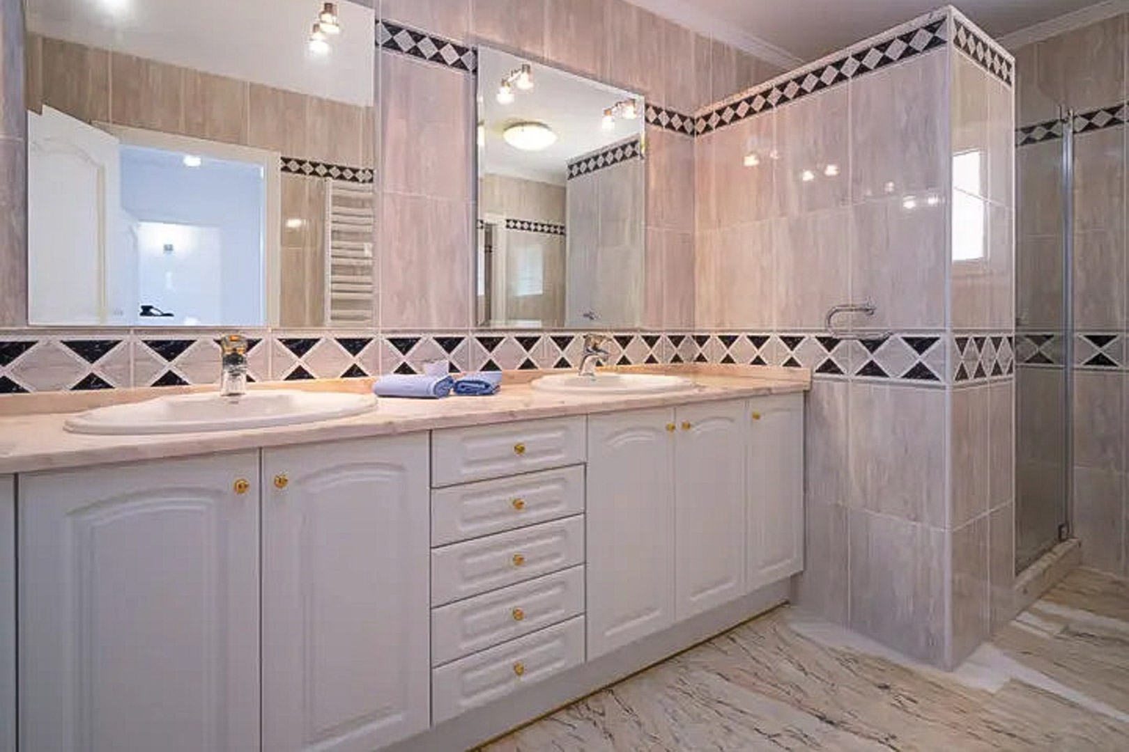 Bathroom featuring a double vanity with white cabinetry, marble countertops, and twin mirrors. The space includes decorative diamond-patterned tiling and a walk-in shower.