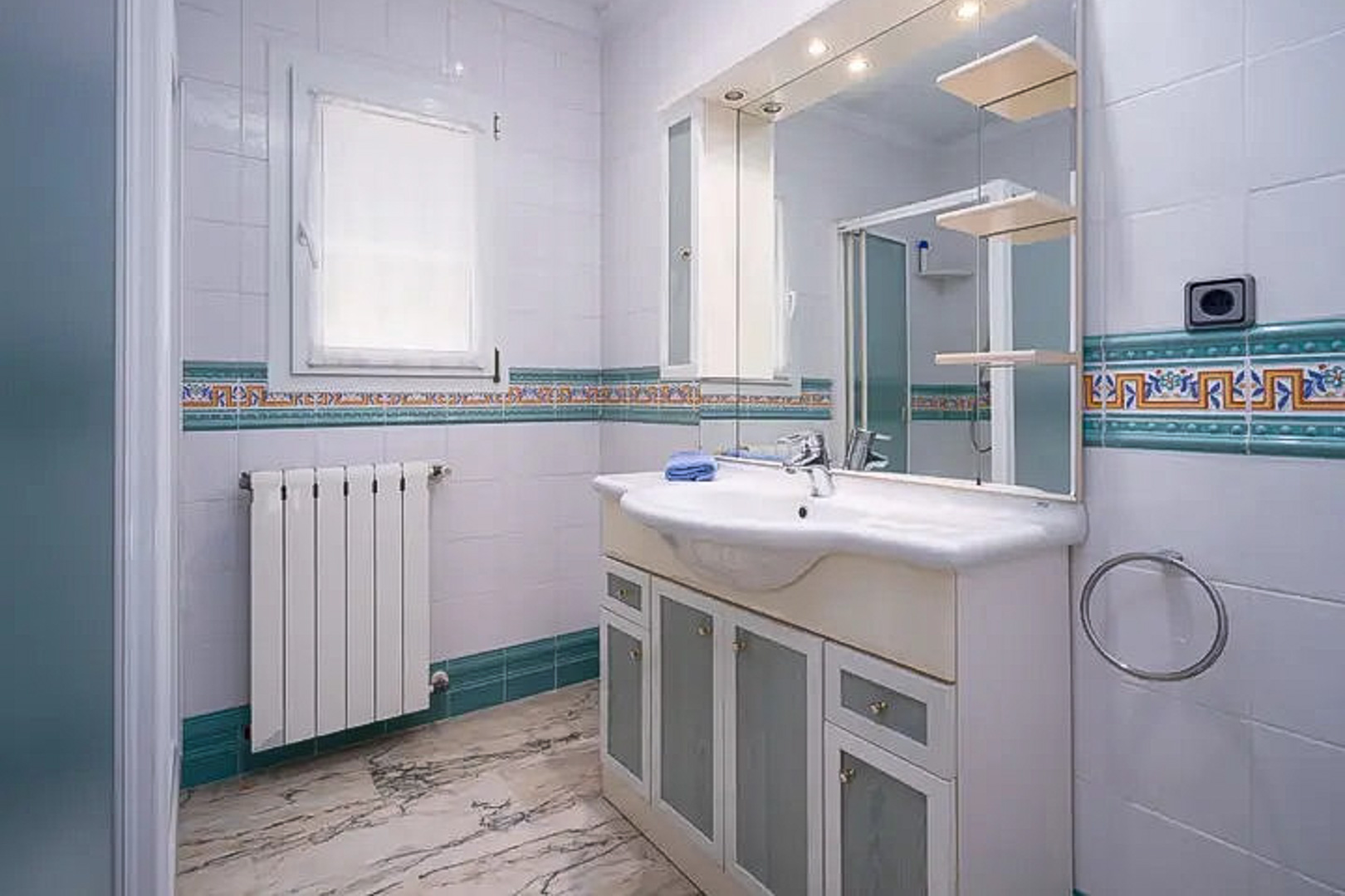 Bathroom featuring white wall tiles with a Mediterranean border, a vanity unit with frosted glass doors, a large illuminated mirror, and marble-effect flooring.