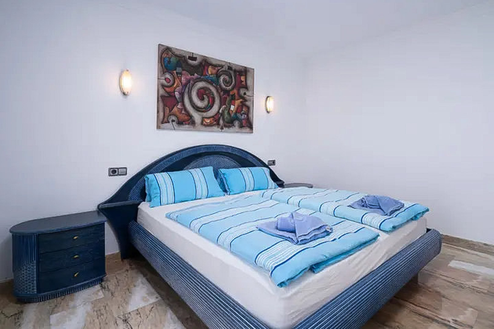Bedroom featuring a blue wicker headboard and matching nightstand. The space includes polished marble flooring, symmetrical wall sconces, and abstract artwork.