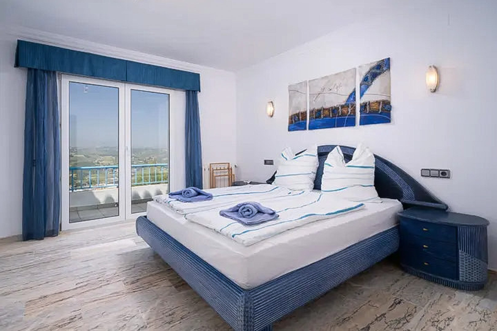 Bedroom featuring marble flooring, a blue-themed double bed, and large glass doors opening onto a private balcony with coastal views.