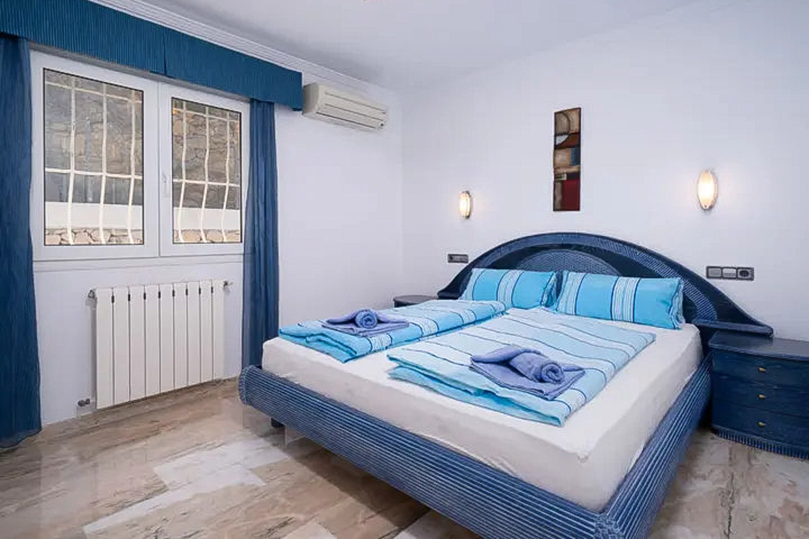 Bedroom featuring polished marble flooring, a blue-themed double bed, split air conditioning, and a radiator positioned under a barred window.