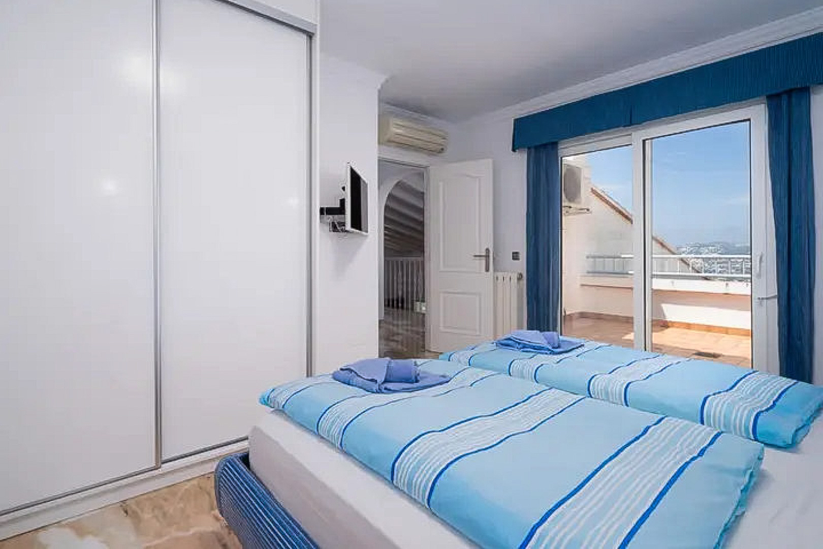 Bedroom featuring twin beds, a large white sliding wardrobe, and direct access to a private terrace. Equipped with air conditioning, a wall-mounted TV, and tiled flooring.