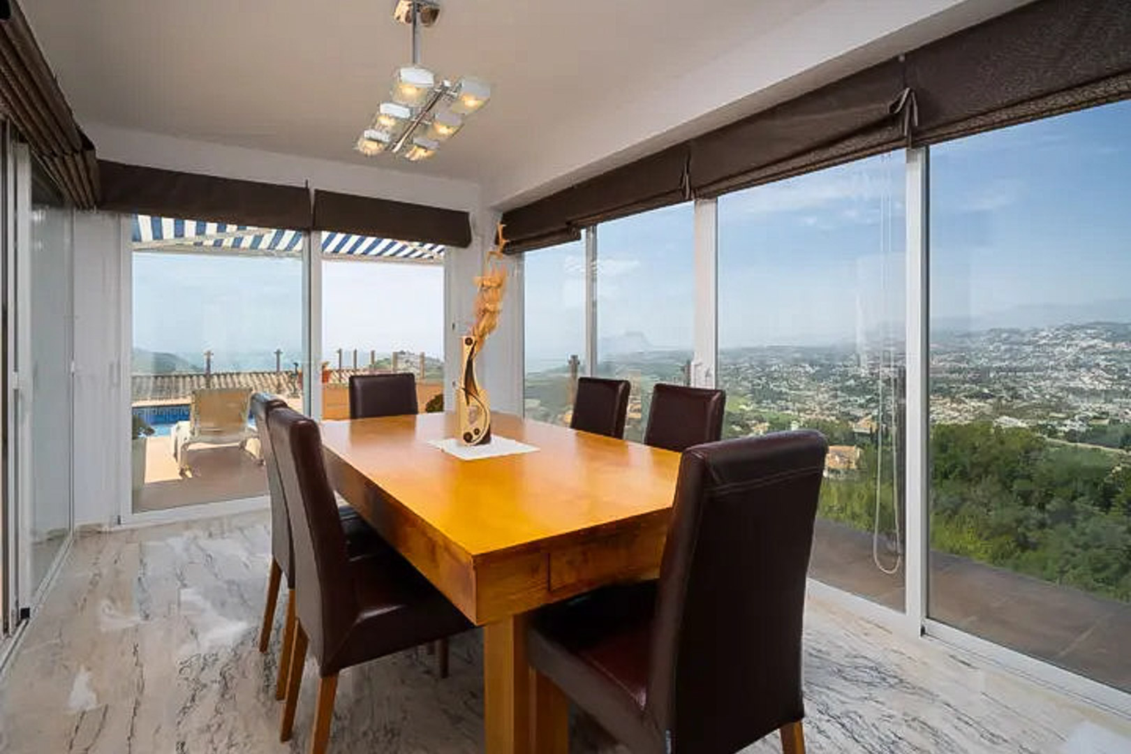 Bright dining area featuring a solid wood table and leather chairs, enclosed by large glass sliders offering panoramic Mediterranean views and terrace access.