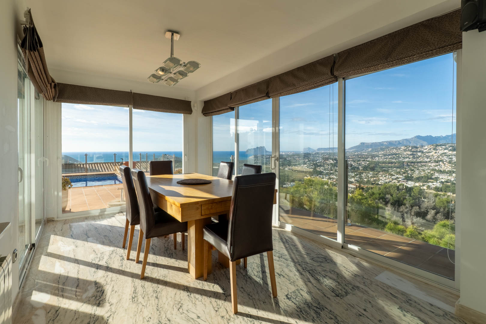 Bright dining area featuring marble flooring and a solid wood table. Large windows provide panoramic views of the Mediterranean Sea, Peñón de Ifach, and the surrounding mountains.